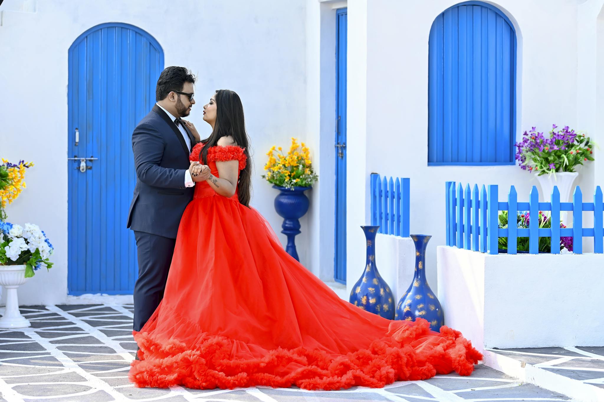 A couple elegantly dressed in a red gown and suit dances lovingly outdoors with vibrant blue accents.